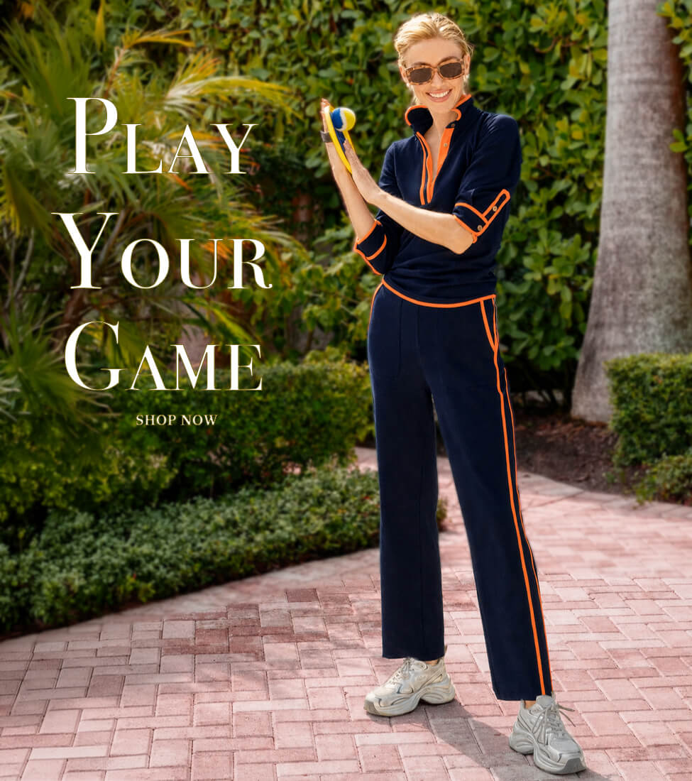 Gretchen Scott Designs model wearing a navy blue athletic tracksuit with orange trim, white sneakers, and sunglasses, holding a pickleball paddle on a garden patio. Text reads: “Play Your Game” and “Shop Now.”