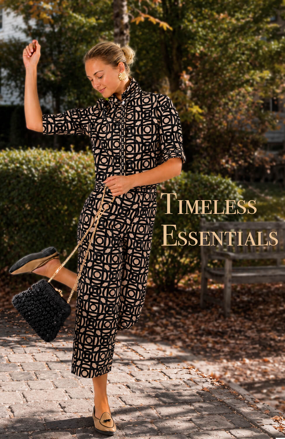 Gretchen Scott Designs model wearing a black and beige geometric-print women’s dress, carrying a black handbag on a garden path. Text reads: “Timeless Essentials.”