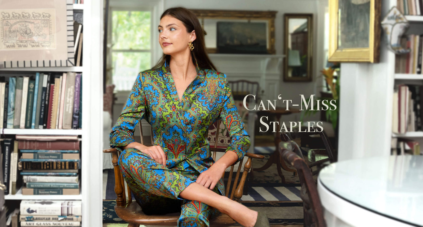 Gretchen Scott Designs model wearing a colorful paisley-print women’s dress, seated in an elegant home library. Text reads: “Can’t-Miss Staples.”