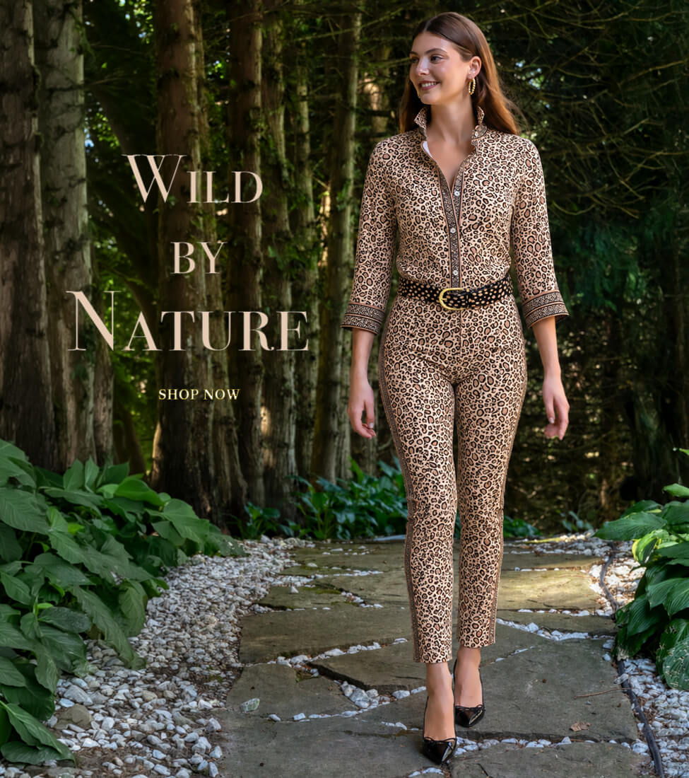 Gretchen Scott Designs model wearing a leopard-print women’s jumpsuit with a black belt and black heels, walking along a garden path surrounded by trees and greenery. Text reads: “Wild By Nature” and “Shop Now.”