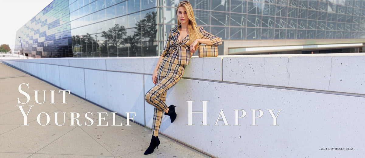 Gretchen Scott Designs Fall/Winter model wearing a tailored plaid women’s suit with black ankle boots, leaning against a modern glass building in New York City. Text reads: "Suit Yourself Happy."